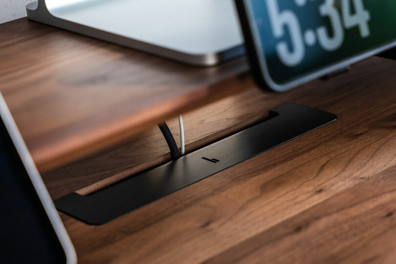 A black Balolo cable management tray mounted beneath the wooden stand holding several cords.
