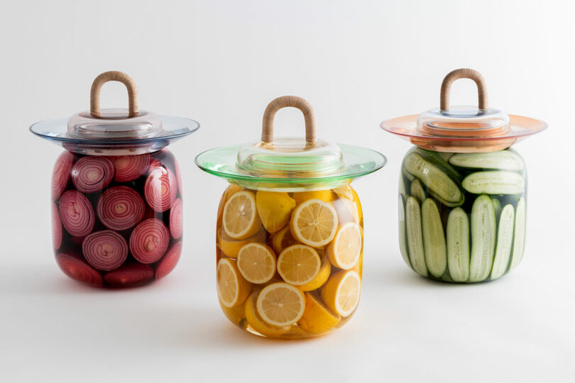 Three glass jars with rustic rope handles containing pickled onions, lemons, and cucumbers.