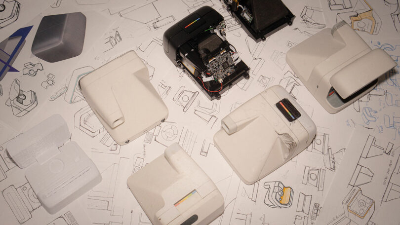 A collection of prototype camera casings and internal components on top of technical design sketches.