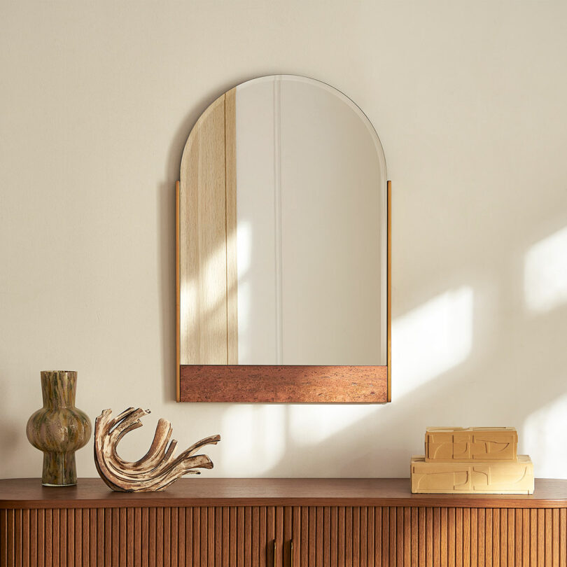 A wooden sideboard styled with a green vase, books, and an arched mirror, creating a sophisticated home vignette.