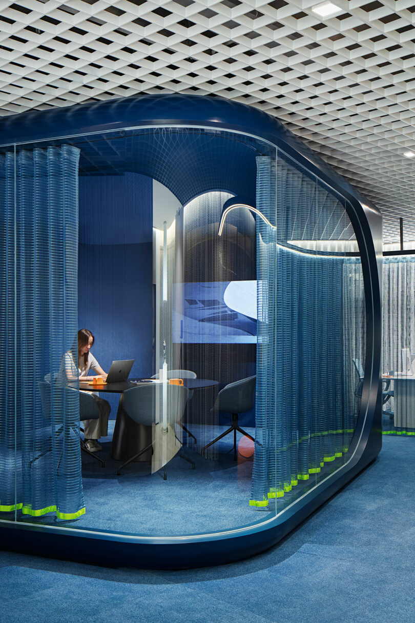 A person working on a laptop inside a glass-walled office pod with blue curtains.