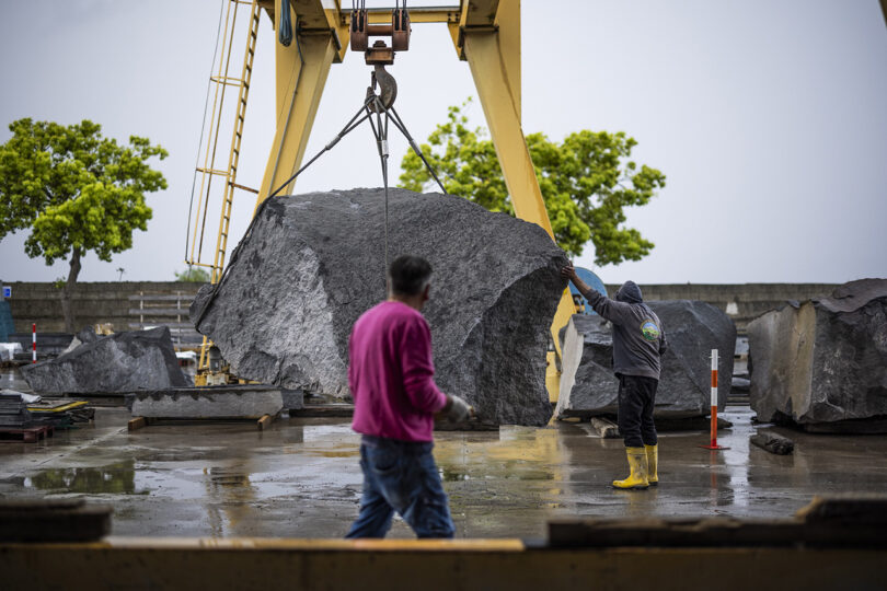 Workers at an industrial site managing a large piece of natural stone suspended by a crane.