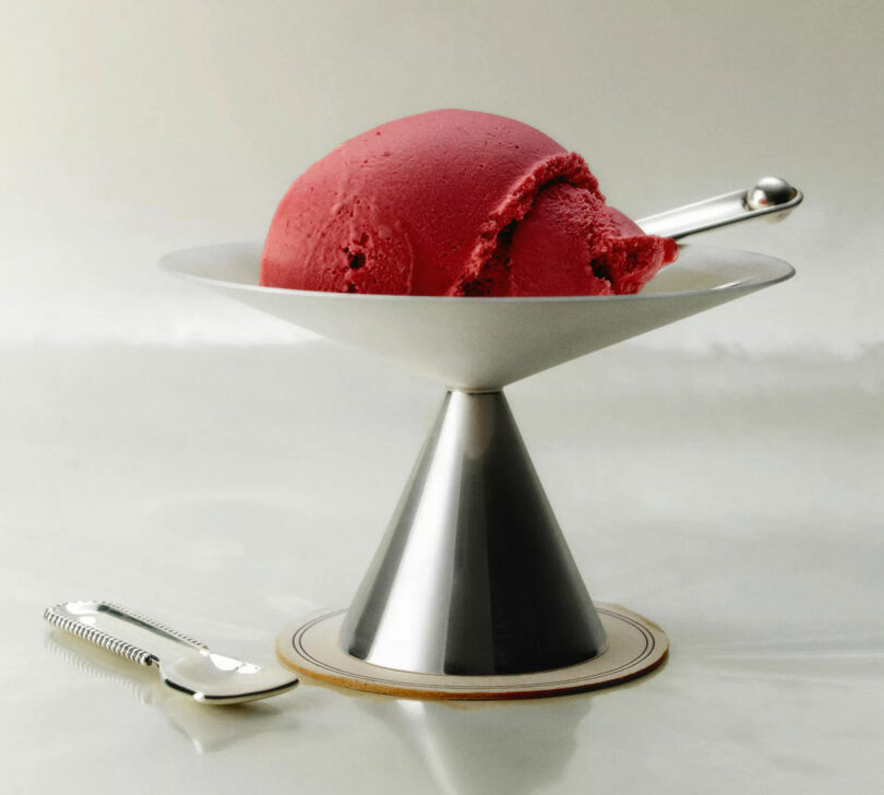 A scoop of raspberry ice cream in a sleek metal dessert dish with a matching spoon on a blue coaster.