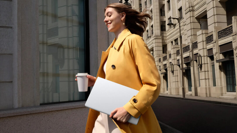 A woman walking through an urban environment carrying a slim laptop and coffee, representing the mobile professional.