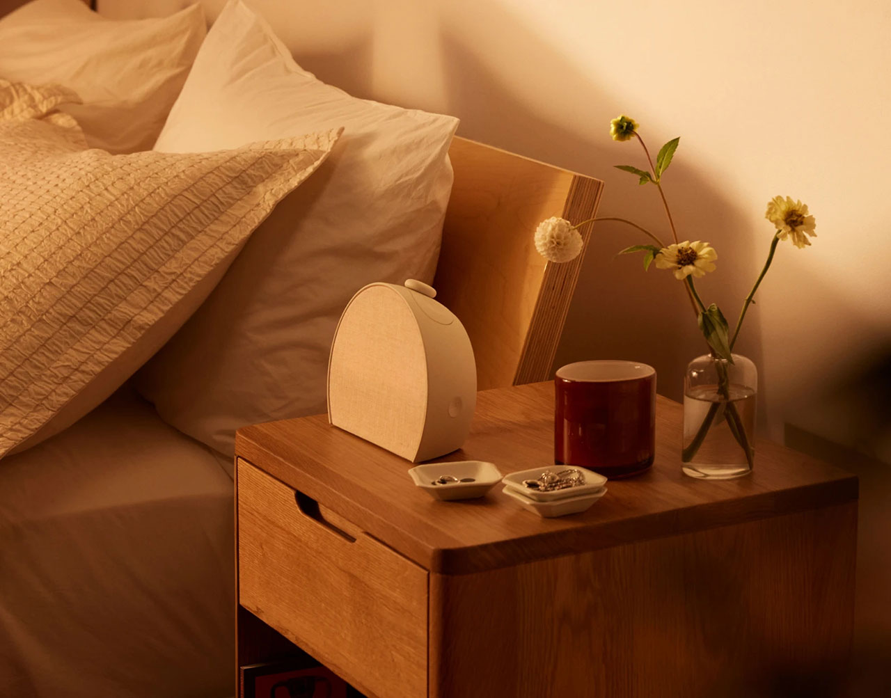 A bedside table featuring a white noise machine, a mug, and a small vase of flowers.