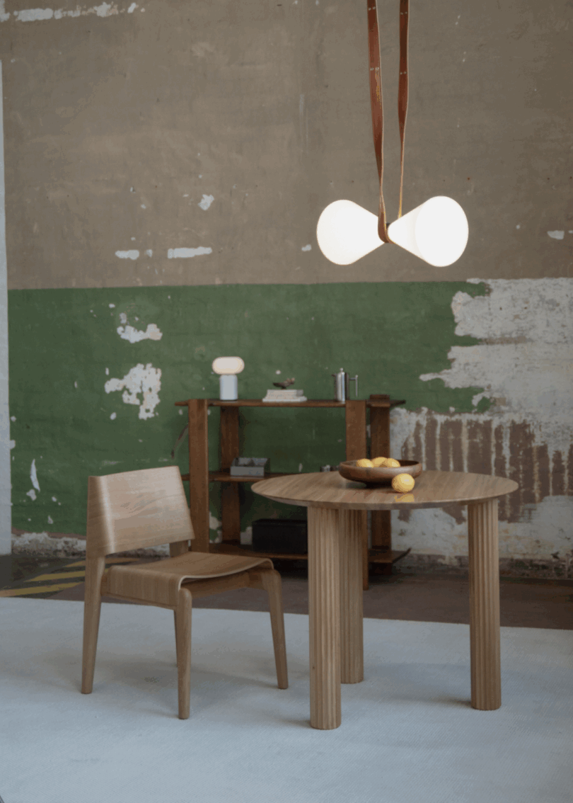 A wooden chair and round table in a minimalist room with a white rug and textured wall.