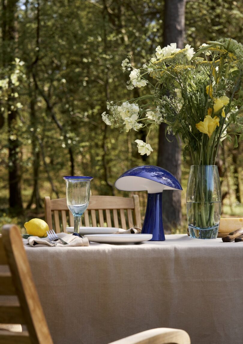 A blue Rumee lamp on a wooden outdoor table set for a meal in a forest setting.