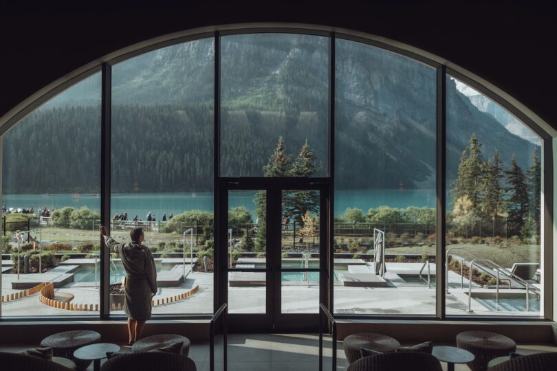 A person in a white robe looking out a window at the mountains and lake at Fairmont Chateau Lake Louise.