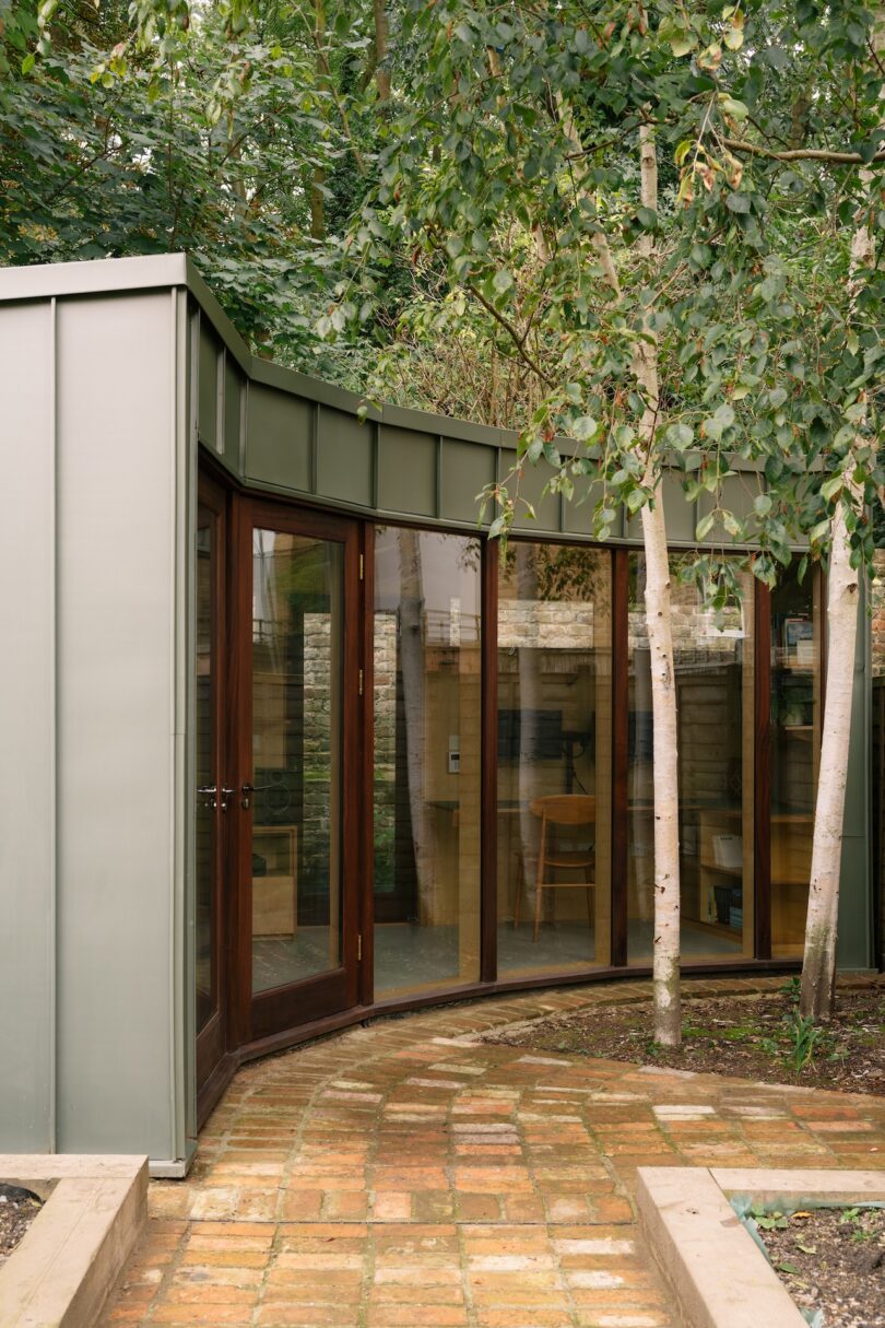 A glass-walled garden room with a metal roof and wooden door under a canopy of trees.
