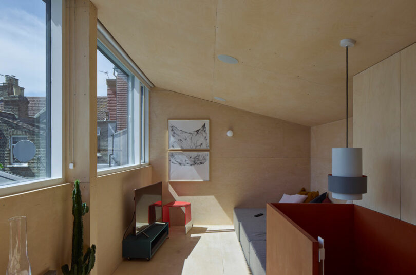 A minimalist loft room with a sloped ceiling, light wood walls, a gray sofa, and a cactus near a large window.