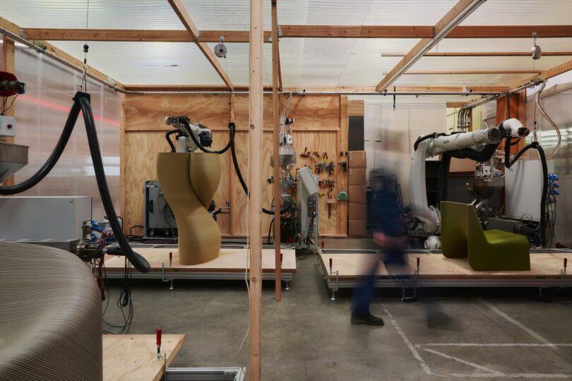 A robotic arm in a workshop 3D-printing a large sculptural furniture piece from recycled plastic.