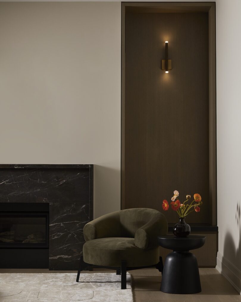 A green armchair next to a dark marble fireplace and a wall sconce.