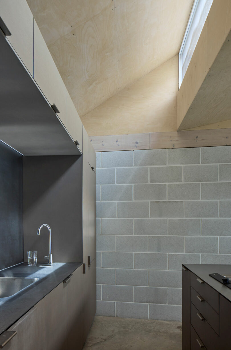 A modern kitchen with concrete block walls, light wood cabinetry, and a slanted ceiling with a skylight.