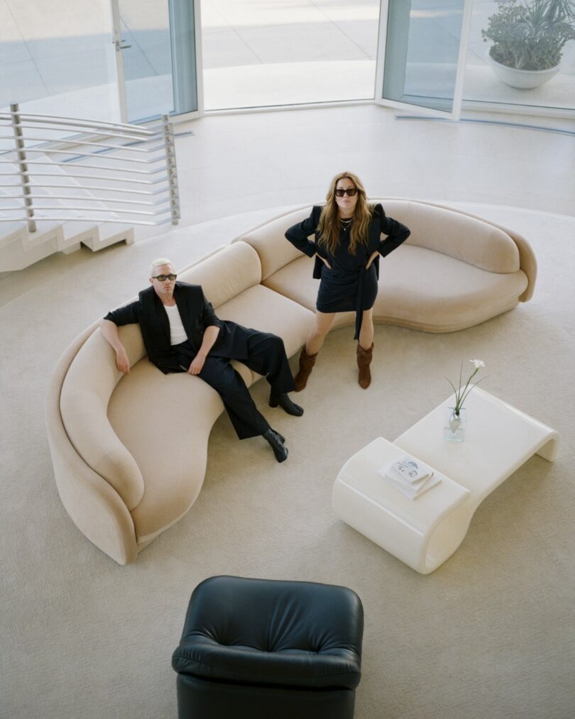 Editorial photo of two people in sunglasses posing on a curved beige Poet Sofa in a modern minimalist living room.