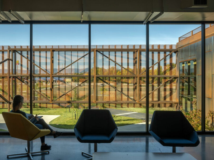 A person sitting in a yellow chair reading near large windows with views of rusted metal structural beams.