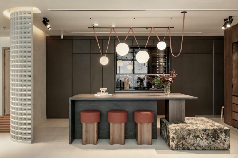 Contemporary office kitchen with a stone island, red stools, and designer lighting.