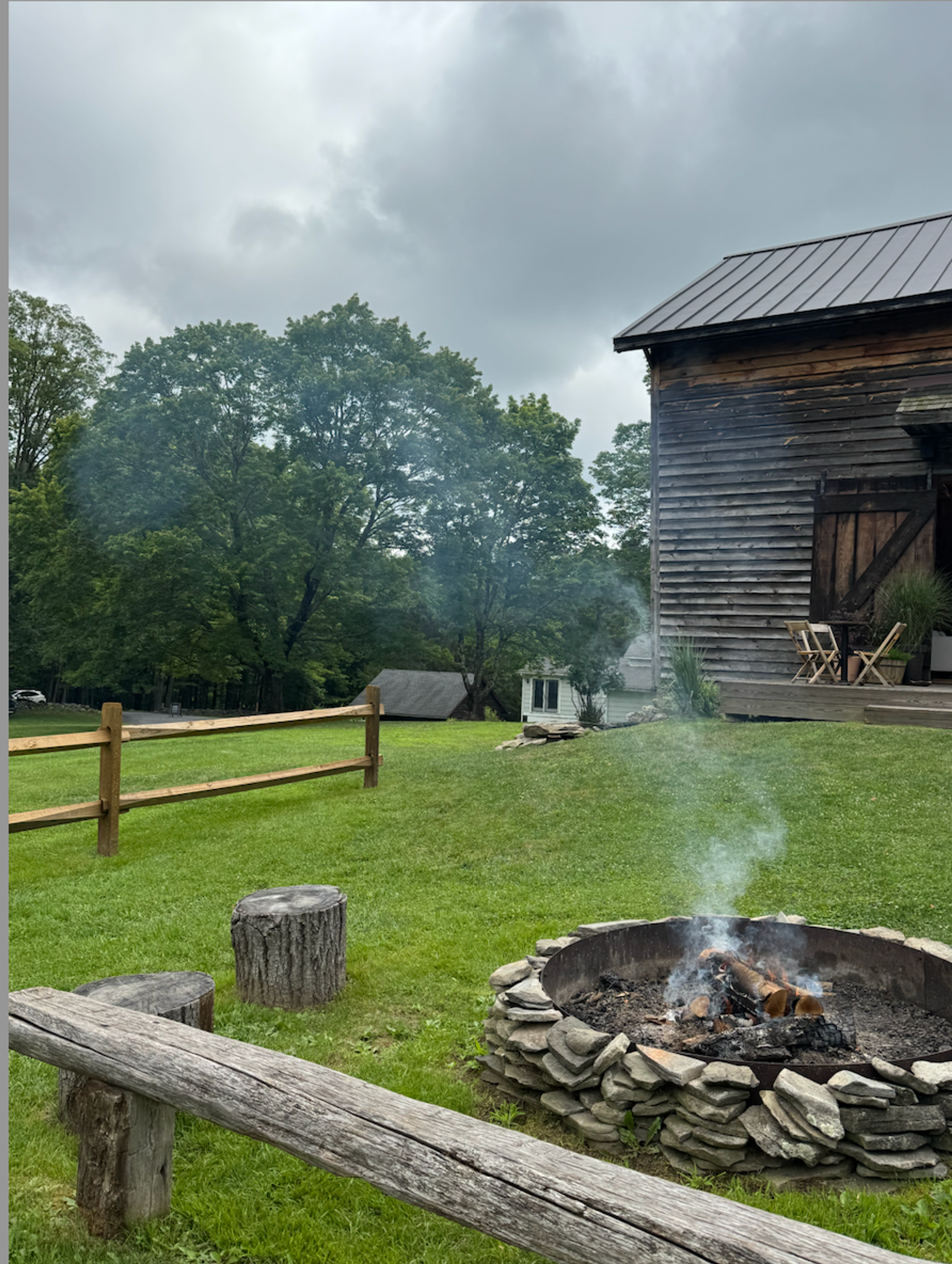 A barn in an open field at dusk with the warm glow of a campfire nearby.
