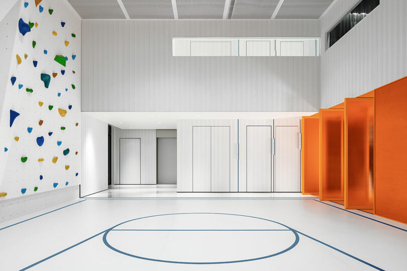 A modern indoor gym featuring a blue basketball court and a colorful rock climbing wall.
