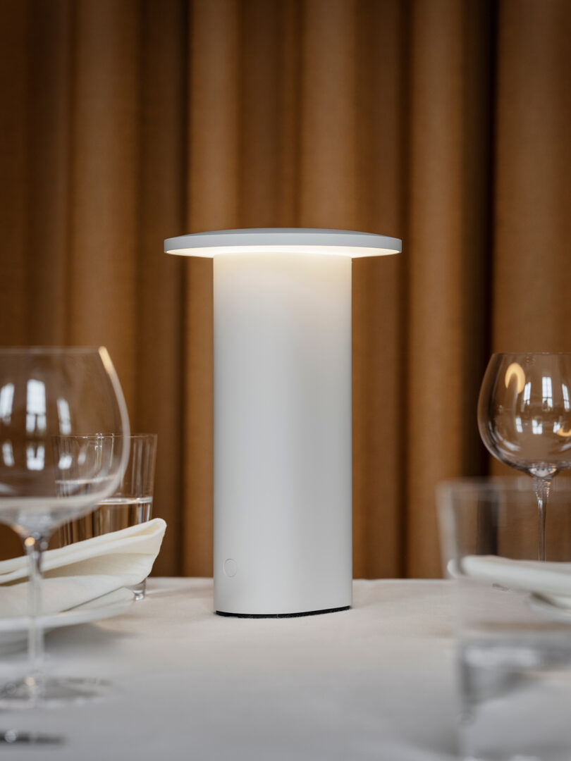 A white cylindrical table lamp glowing softly on a dinner table with glassware and napkins.