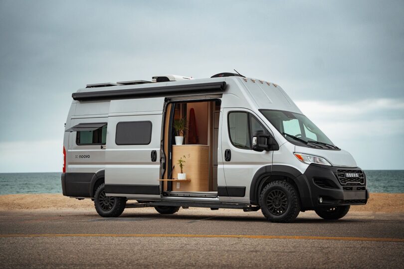 A silver camper van with its side door open to reveal a minimalist wood-paneled interior.