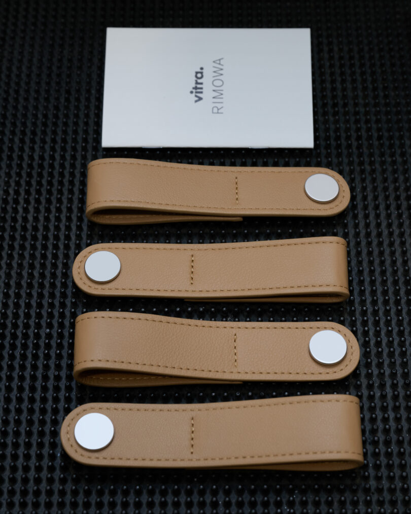 Four tan leather straps with silver snaps arranged neatly under a Vitra RIMOWA collaboration card.