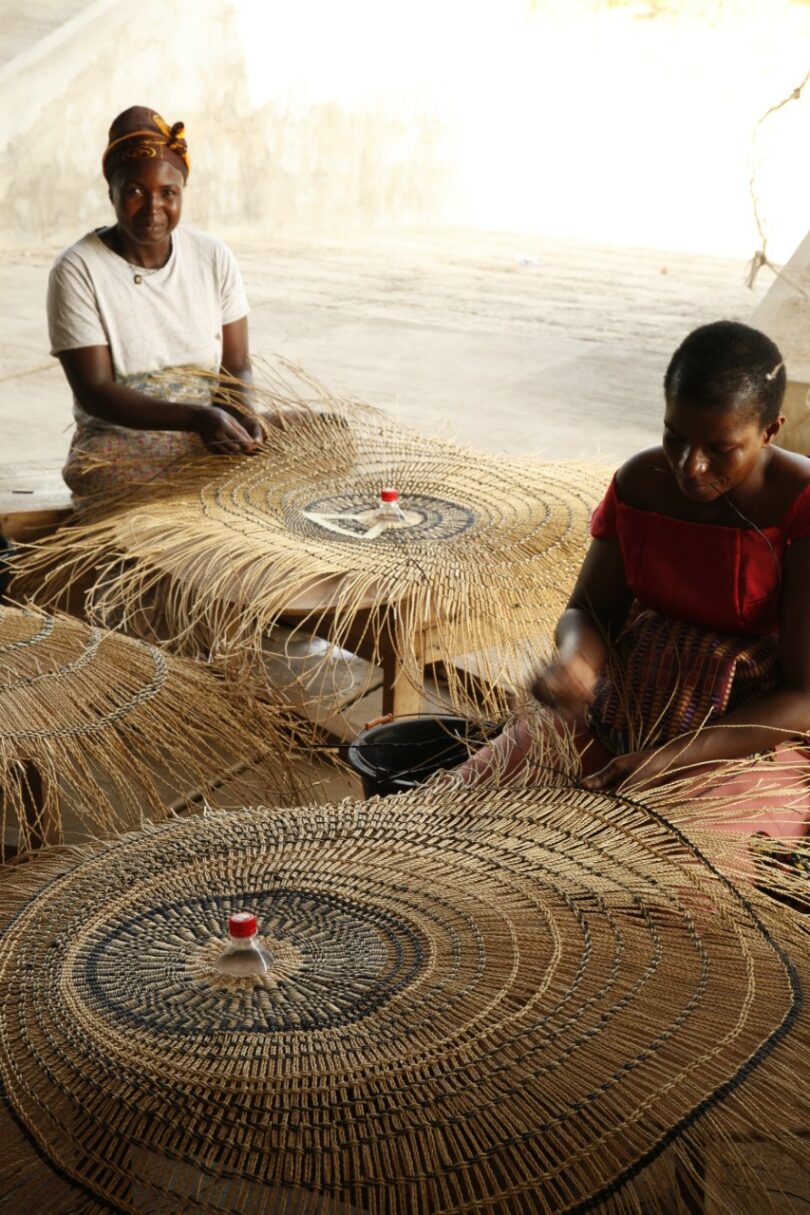 Two Ghanaian women sitting on the floor weaving large circular fiber baskets anchored by red plastic bottle caps.