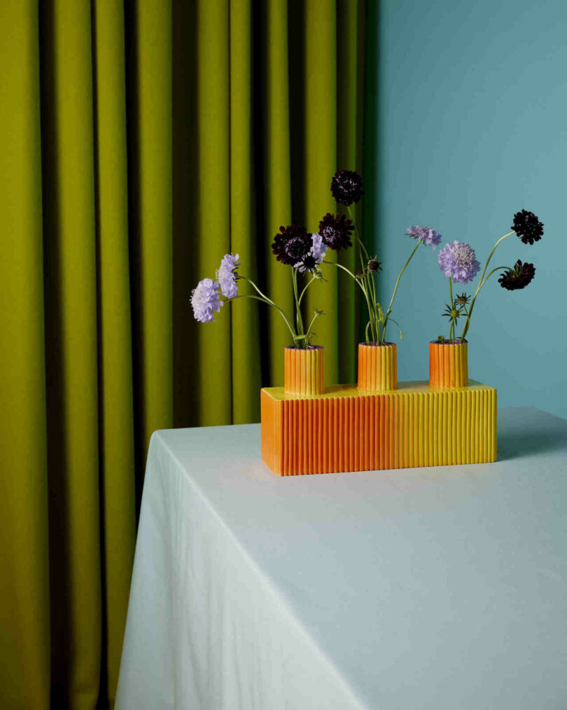 A triple-spout orange ceramic vase holding purple flowers against a teal and blue background.