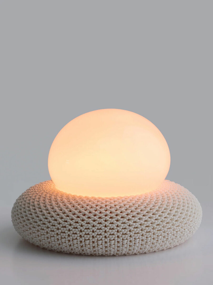 A round, glowing white lamp resting on a 3D-printed base that resembles a knitted textile texture.