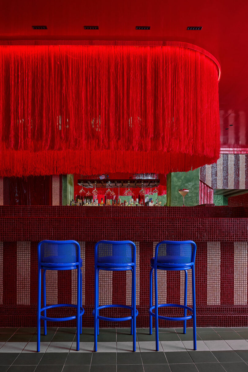 A bar interior featuring a red tiled counter, blue stools, and a dramatic red fringe ceiling installation.