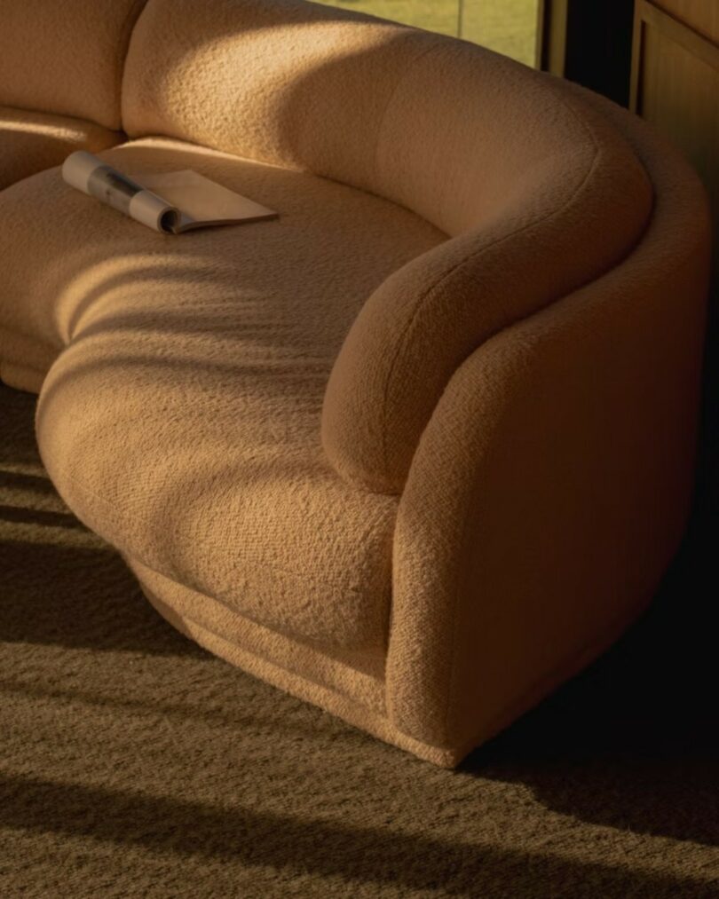 Close-up of a beige curved sofa in direct sunlight with a newspaper resting on the textured fabric.