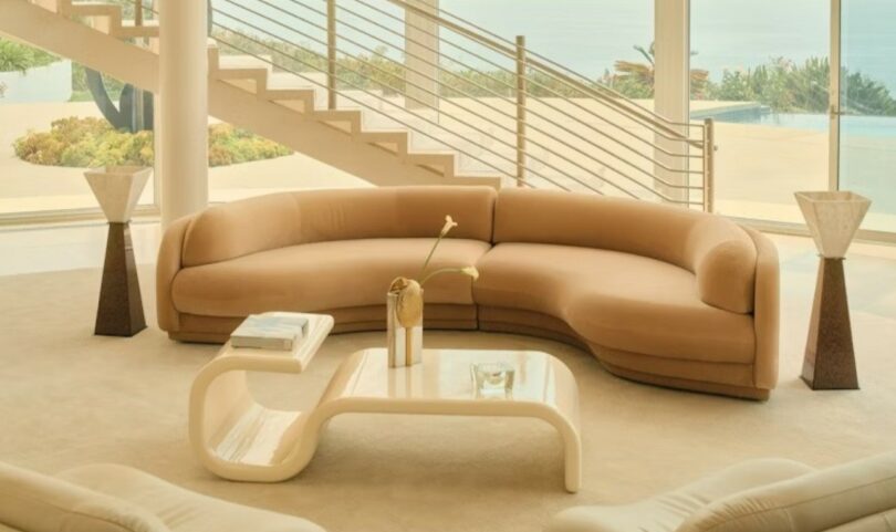 A luxury living room featuring a curved beige sofa and sculptural coffee table with a view of a pool and staircase.