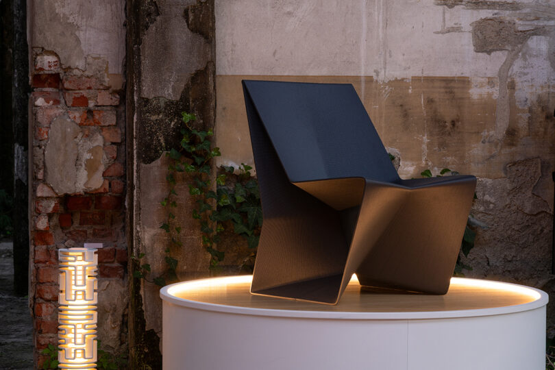 A black angular 3D-printed chair with geometric facets on a glowing white platform.