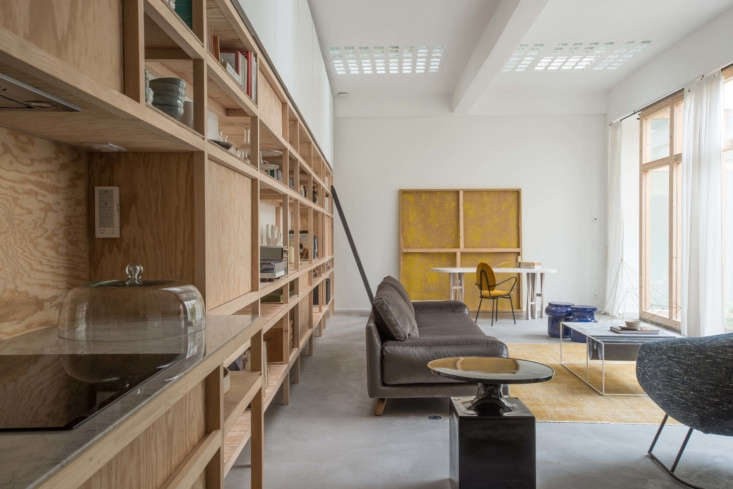 Custom pine plywood shelving extending along a long living room wall with original glass ceiling tiles above.