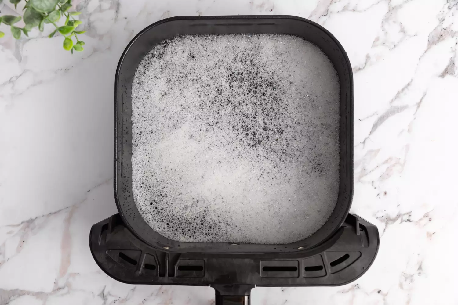 A black air fryer basket soaking in soapy water on a modern marble countertop.