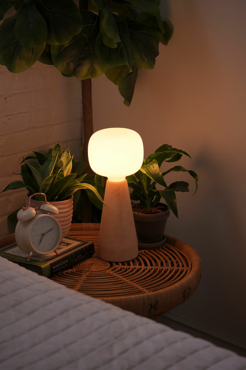 The Aura lamp on a wooden nightstand next to a bed with books and indoor plants.