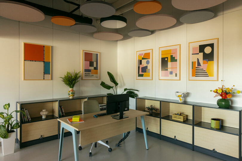A modern wooden office desk with a computer and ergonomic chair positioned against a wall with geometric artwork.