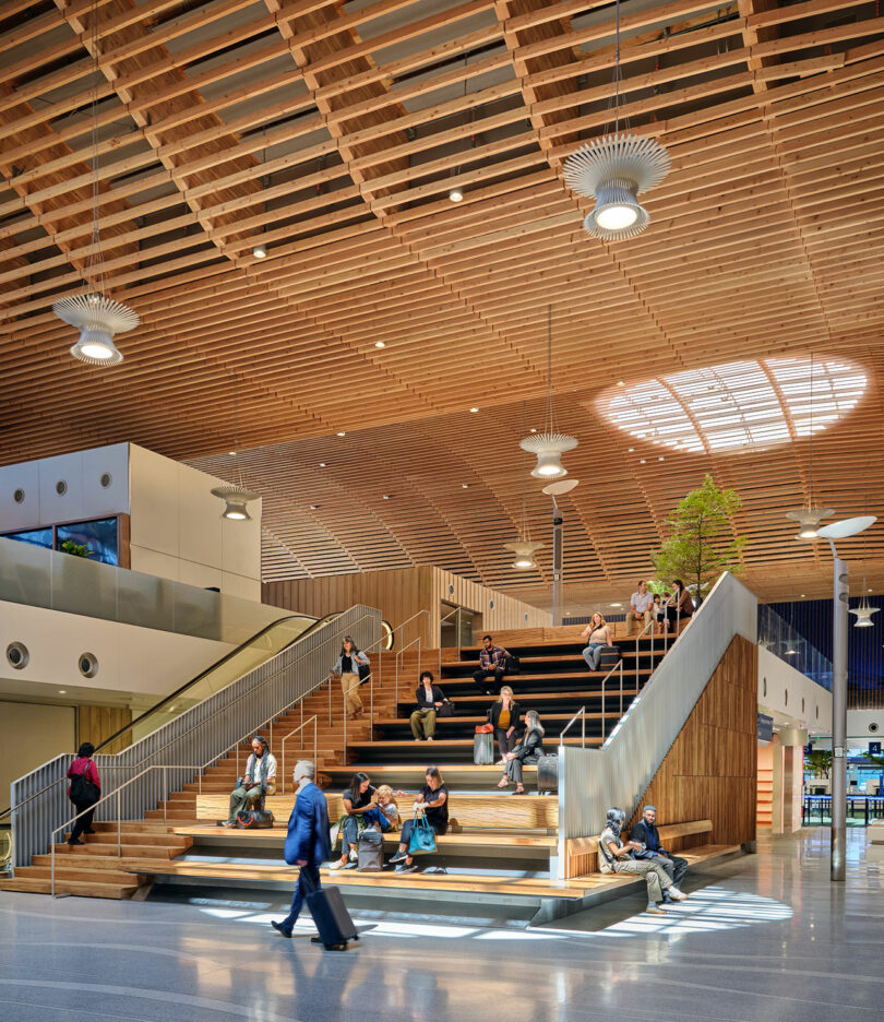 People sitting on a large wooden indoor staircase under a modern skylight.