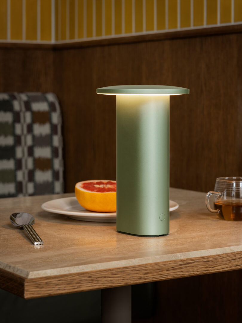A green cylindrical table lamp illuminated on a table with a grapefruit and tea.