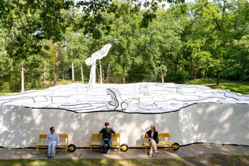 Three people sitting on yellow benches in a park setting in front of a large outdoor mural.