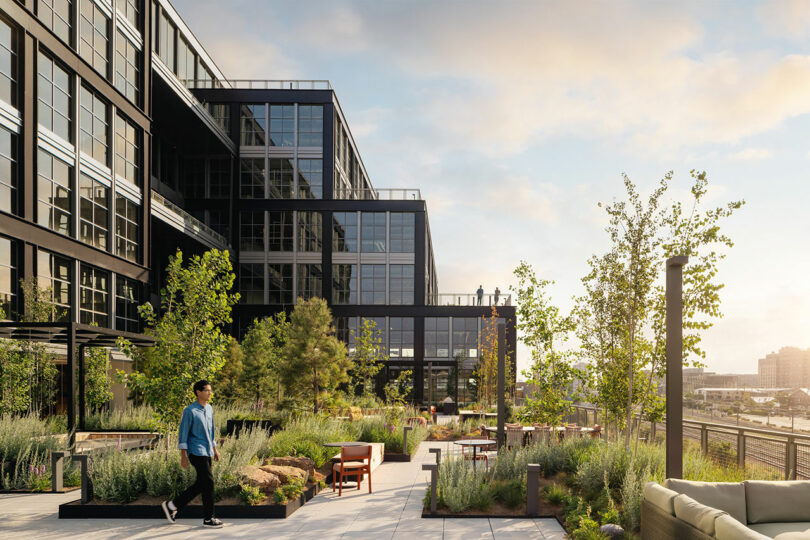 A person walks along a landscaped rooftop terrace with seating areas and lush greenery at Steel House.