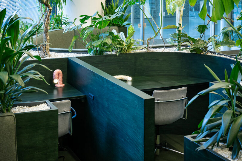 A focused workspace with dark wood desks, grey chairs, and worm-shaped lamps separated by plant dividers.