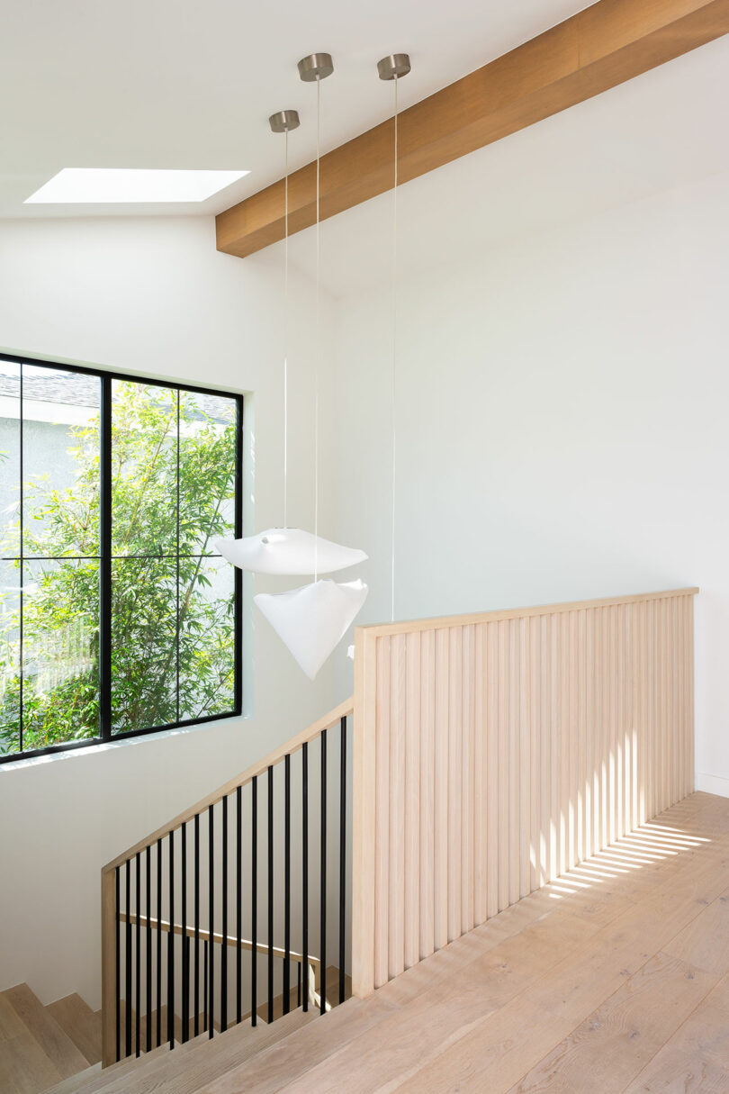 Modern stairway with light wood railing and vertical black balusters, illuminated by geometric pendant lights and a large window.
