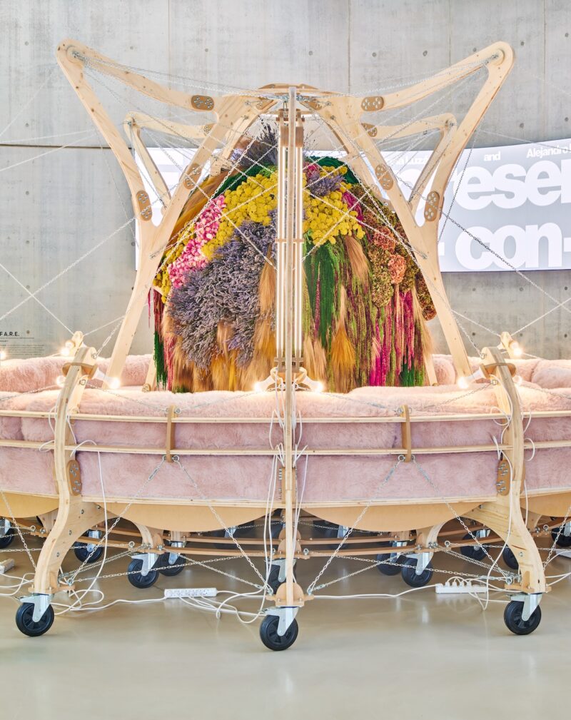 A close-up of a pink cushioned seat on a wooden wheeled base supporting a central bouquet of colorful flora.