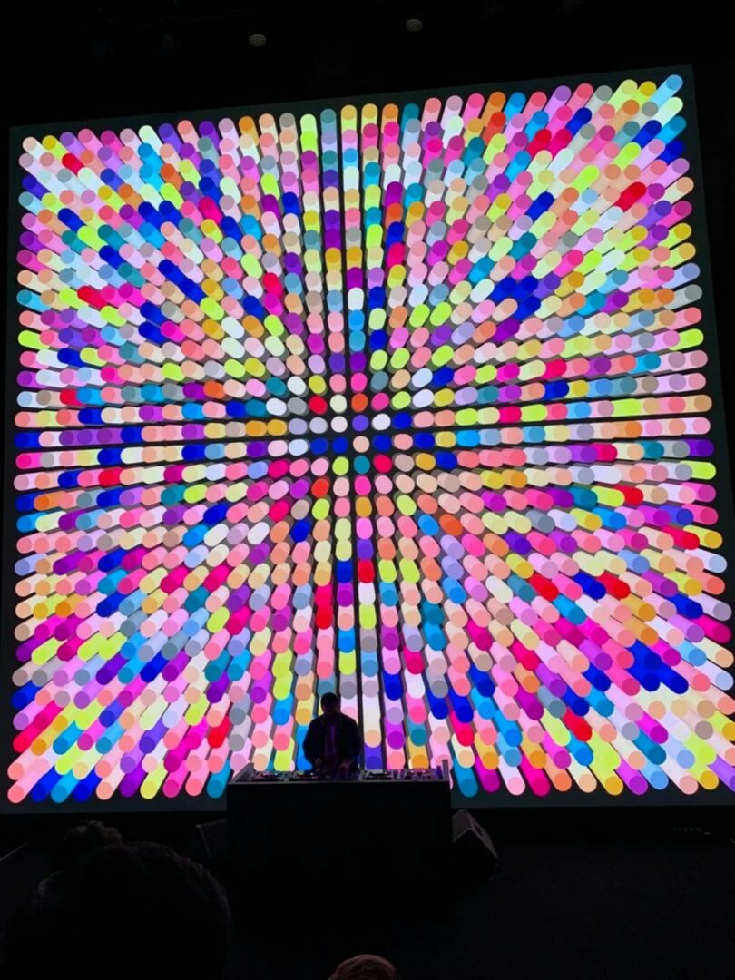 A person at a DJ booth performing in front of a massive circular generative art display.