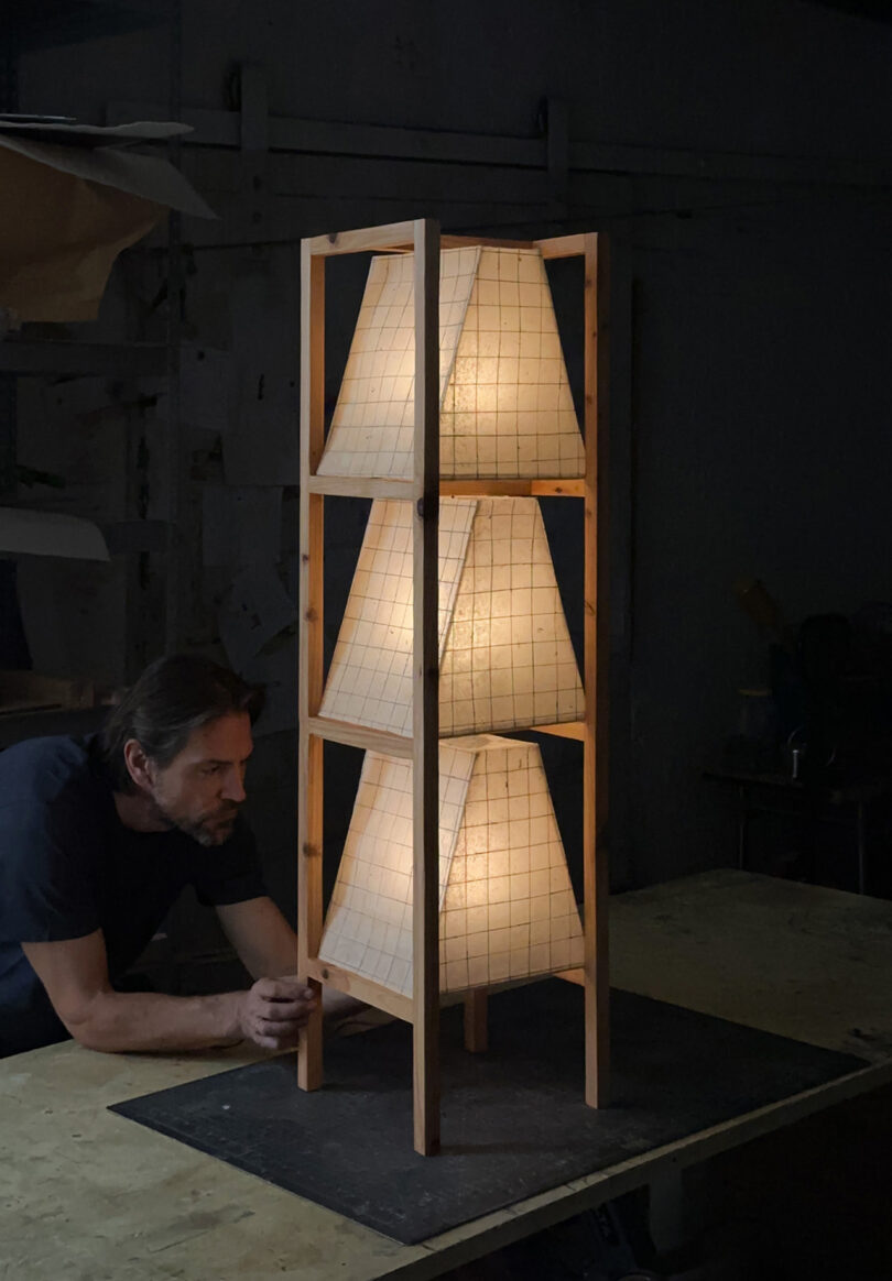 A designer adjusting a large wooden floor lamp frame with three stacked grid-patterned paper shades.