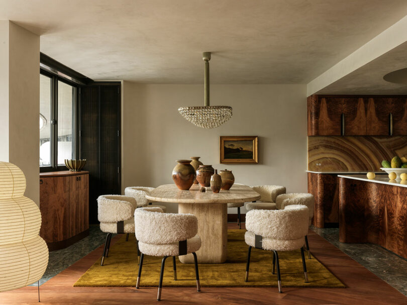 A dining room with a round stone table, textured white chairs, and a large crystal chandelier.