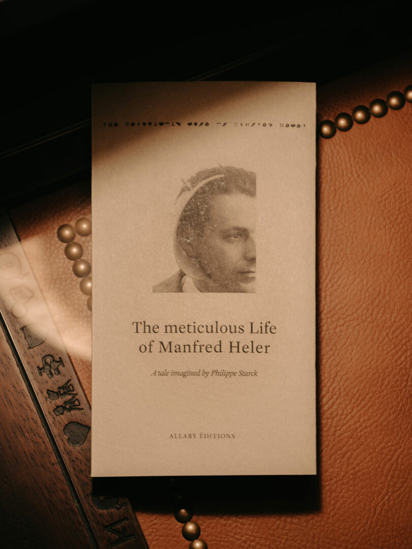 A sepia-toned book titled 'The Meticulous Life of Manfred Heler' lying on a leather surface.