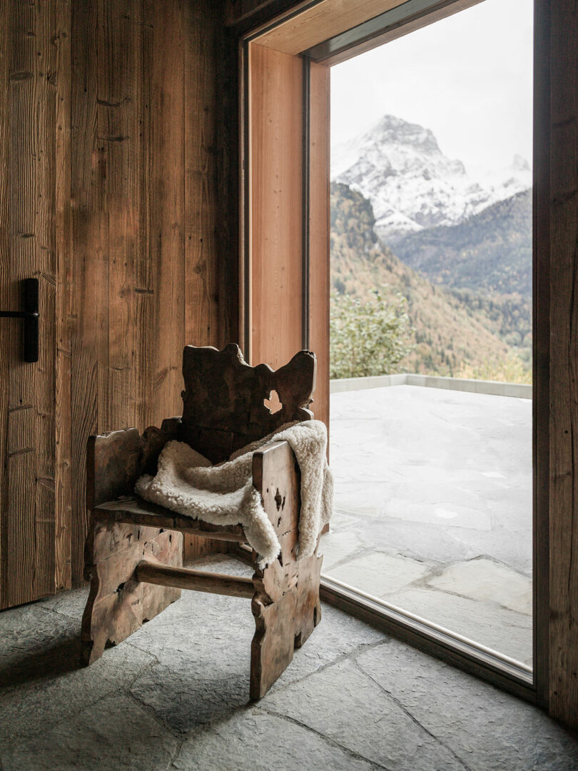A rustic chair by a large window with a view of alpine peaks and a stone patio.