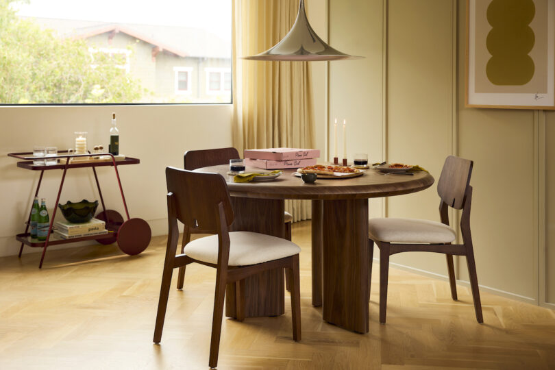 A modern dining room featuring a round wooden table and a bar cart positioned near a bright window.