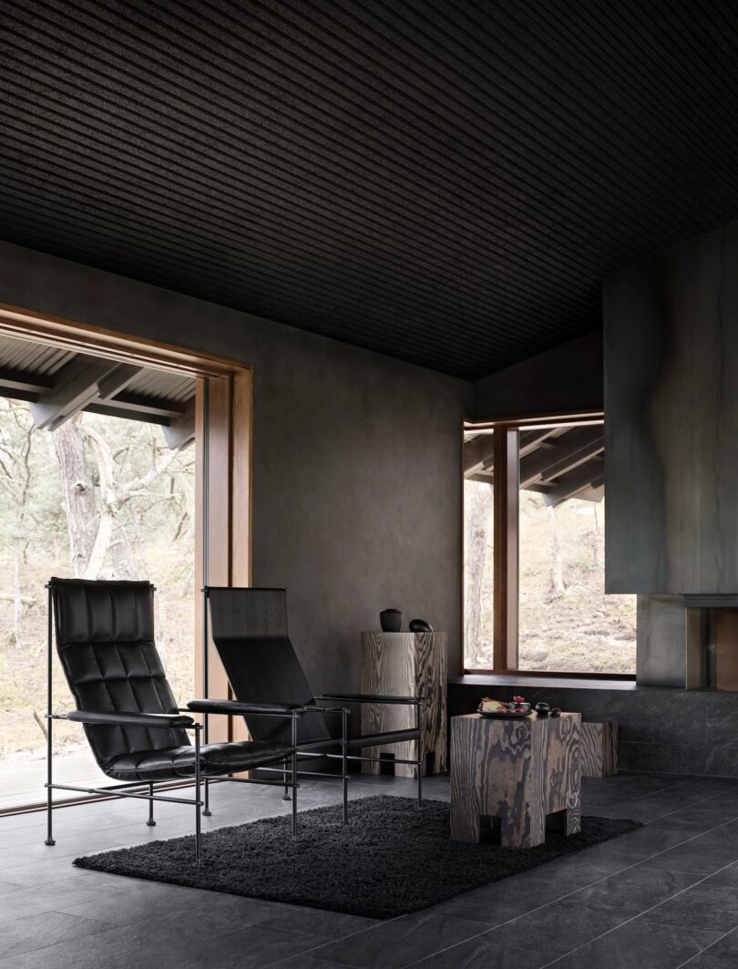 Modern minimalist living room with dark tones, two leather chairs, and large windows overlooking trees.